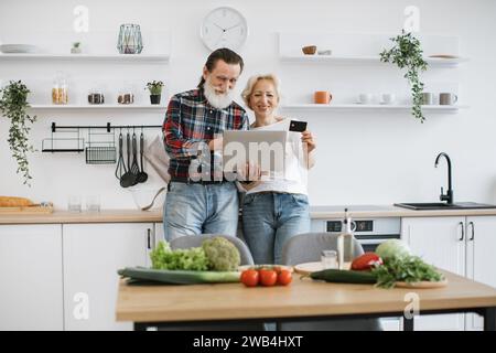 Old Caucasian couple using laptop and credit card to buy food products. Stock Photo