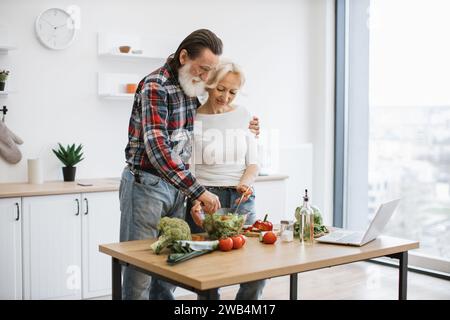 Caucasian old couple spends free time cooking breakfast in modern light kitchen. Stock Photo