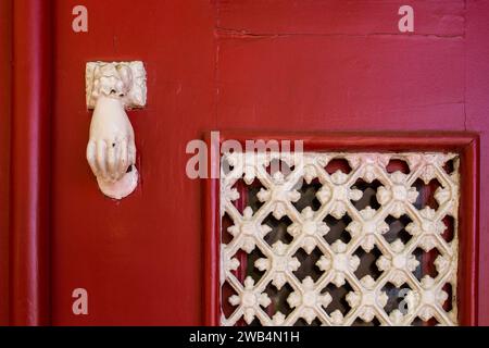 Bright red painted wooden entrance door with a white knocker in a shape of a hand and an ornamental white grating in the window. Porto, Portugal. Stock Photo