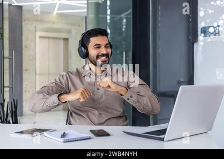 A young Indian man works in the office. He sits at his desk with headphones in front of his laptop and dances during the break, rests, rejoices. Stock Photo