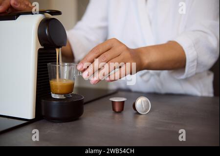 Close-up of a housewife's hands using a capsule coffee machine preparing freshly brewed espresso coffee, standing at the kitchen counter in cozy home. Stock Photo