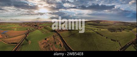 Panoramic aerial view of a scenic landscape with rolling hills, fields, and a winding road under a dramatic sky at sunset. Stock Photo