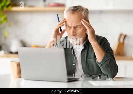 Portrait of stressed senior man using laptop in kitchen interior Stock Photo