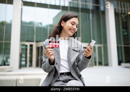 Young businesswoman shopping online making payment on phone sitting outside Stock Photo