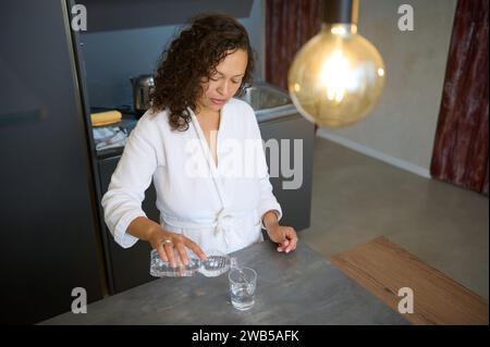 Overhead view of attractive young woman in white bathrobe, standing at kitchen counter in modern minimalist home interior, pouring some water into a d Stock Photo