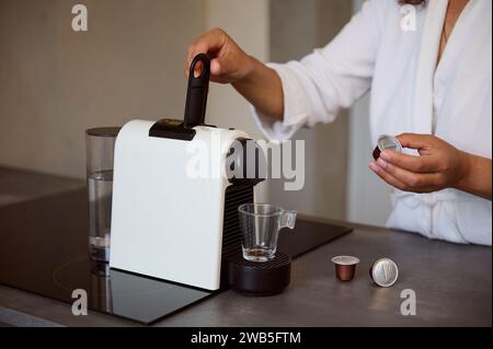 Closeup of a young woman in white waffle bathrobe, preparing freshly brewed coffee in the morning, using a capsule coffee machine. Morning routine. Pe Stock Photo