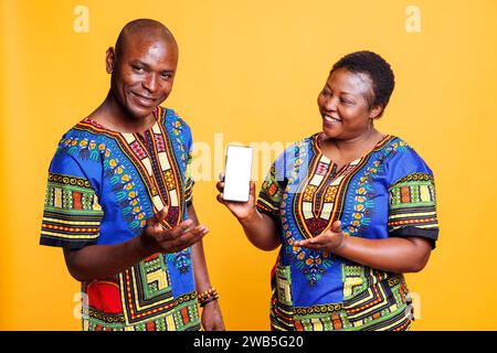 Smiling black couple wearing ethnic closes showing smartphone with white blank screen mockup. Cheerful man and woman presenting mobile phone with empty touchscreen for app advertising Stock Photo