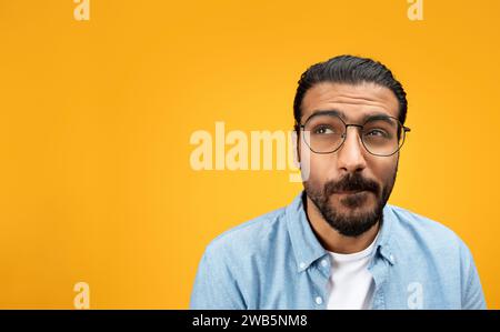 Pensive indian millennial man in glasses, look at free space, think, isolated on orange background Stock Photo