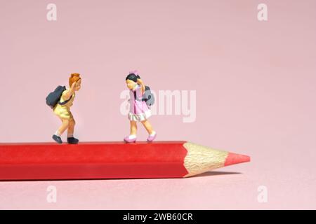 Miniature tiny people toys photography. Two kids standing above red pencil. Isolated on pink background. Image photo Stock Photo