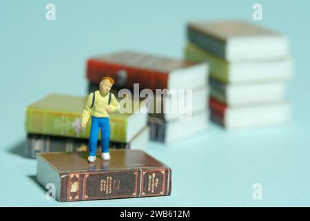 Miniature tiny people toy photography. A boy with school backpack standing above book stair. Isolated on blue background. Image photo Stock Photo
