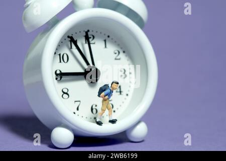 Miniature tiny people toy photography. A boy student with backpack standing on clock. Isolated on purple background. Image photo Stock Photo