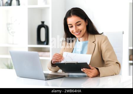 Positive elegant successful indian or arabian business woman, financial manager sitting at workplace in the office, working on a laptop, studying financial reports, glad with results, smiling Stock Photo
