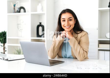 Pretty, positive successful brunette indian or arabian business woman in an elegant suit, ceo, company employee, sitting at a work desk in the modern office, looking at camera and smiling friendly Stock Photo