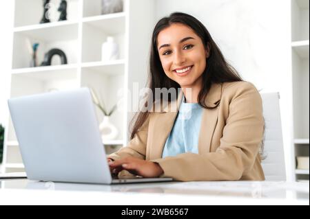 Gorgeous successful brunette indian or arabian business woman, company manager, secretary, sitting at workplace in the modern office, looks at camera and smiling while working on a project on a laptop Stock Photo