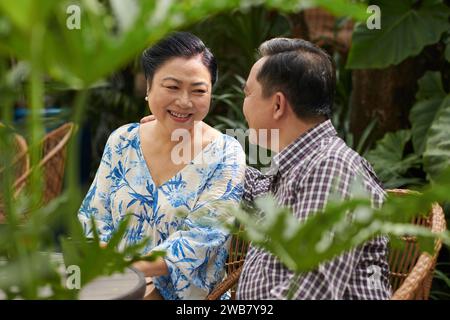 Happy senior Vietnamese man and woman sitting on bench in park and looking at each other Stock Photo