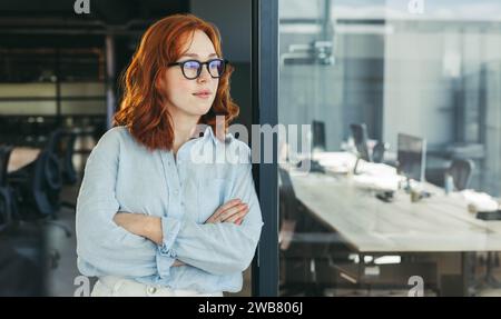 Confident redhead businesswoman in glasses stands in office, arms crossed. Professional and pensive, she reflects on business strategies. Tech-filled Stock Photo