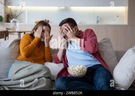 Frightened emotional young couple with popcorn watching horror film, thriller, scary movie at home Stock Photo