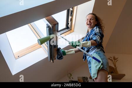 Cheerful young woman has fun wile doing housework cleaning roof window at home. Stock Photo