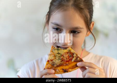 little asia girl keeps slice of pizza in mouth, spreads hands with hesitation, has puzzled facial expression, enjoys nice taste, wants to eat, positiv Stock Photo
