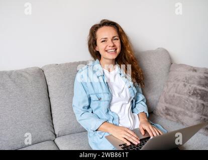 Happy young brunette woman caucasian ethnicity sits on the sofa at home and using her laptop computer. Stock Photo