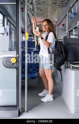 Young urban woman standing on a bus and smiling while using her smartphone. City public transportation and passenger lifestyle. City life. Stock Photo