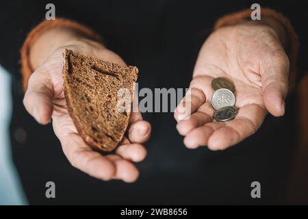Hungry woman holding money and bread on a black background, hands with food close-up. Cash in the dirty hands of a starving poor woman on a dark backg Stock Photo