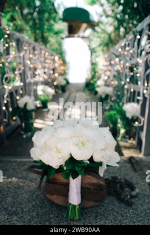 White bridal bouquet placed on a rustic chair along a decorated outdoor wedding aisle surrounded by greenery. Stock Photo