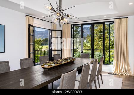 A contemporary wooden dining table surrounded by matching chairs in a bright, airy room illuminated by a large window Stock Photo