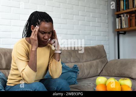 African American woman sitting at home in apartment having issue and headache because of loud noise coming from apartment above. Sick or ill female, n Stock Photo