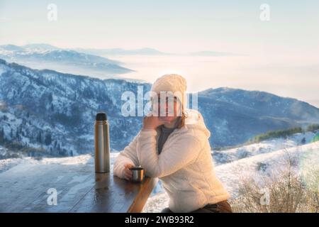 Happy woman drinks tea from a thermos against the backdrop of snow capped mountains in winter Stock Photo