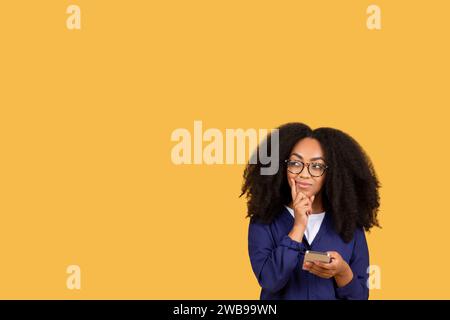 Thoughtful black lady student in glasses holding cellphone, gazing at free space, on yellow background Stock Photo