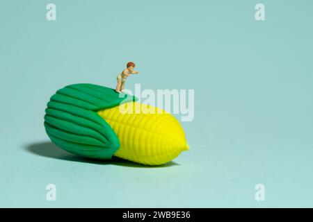 Miniature tiny people toy figure photography. Healthy food. A boy toddler infant feels happy looking at corn. Isolated on blue background. Image photo Stock Photo