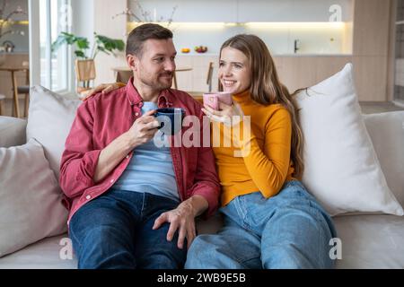 Loving couple man woman spending lazy weekend drink coffee sitting on couch at home, enjoy talking. Stock Photo