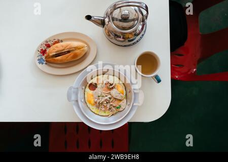 Fried eggs in a small pan with pork and sausages serving on table with Banh mi pork and pickles Vietnamese sandwich and Chinese tea pot. Flat lay food Stock Photo