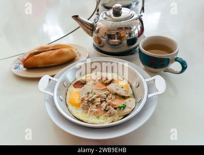 Fried eggs in a small pan with pork and sausages serving on table with Banh mi pork and pickles Vietnamese sandwich and Chinese tea pot. Flat lay food Stock Photo