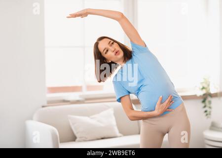 Millennial pregnant woman doing side bends with raised arm indoors Stock Photo