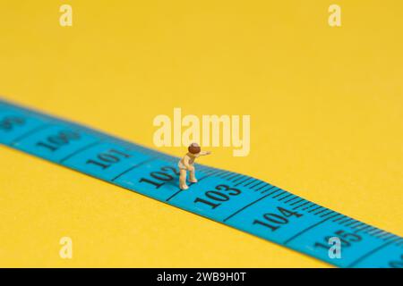 Miniature tiny people toy figure photography. Child growth period concept. A boy toddler walking above blue measuring tape. Isolated on yellow backgro Stock Photo