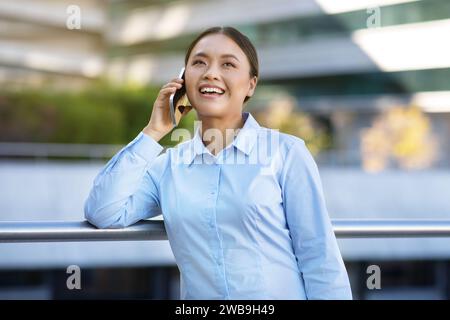 Smiling Korean business lady engaged in phone conversation standing outside Stock Photo