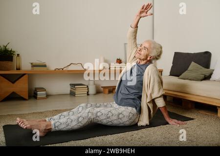 Retired woman in activewear sitting on mat on the floor of bedroom and doing side bends with raised arm while working out after sleep Stock Photo