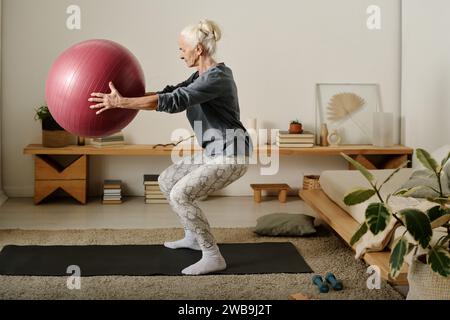 Side view of aged active woman in sportswear holding fitball in front of herself while doing squats on the floor of bedroom in the morning Stock Photo