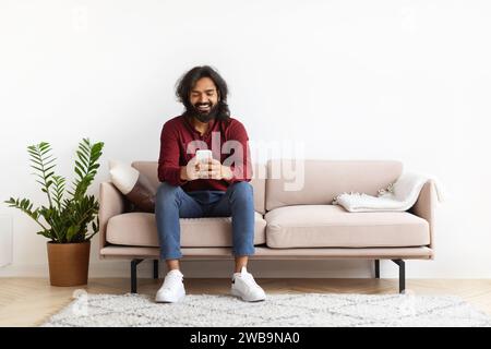Cheerful young indian man sitting on couch, using phone Stock Photo