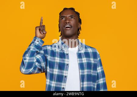 Black man in plaid shirt pointing upwards, looking inspired against yellow background Stock Photo