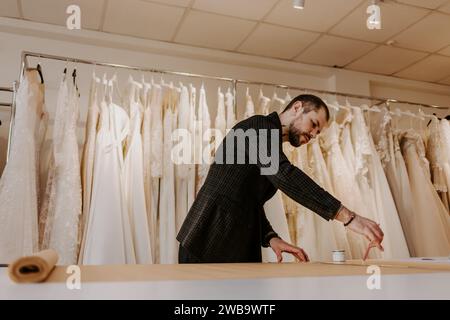 Young serious tailor with beard looking on drawing near wooden table with threads in amazing atelier with antique furniture and mannequin Stock Photo