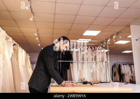 Young serious tailor with beard looking on drawing near wooden table with threads in amazing atelier with antique furniture and mannequin Stock Photo