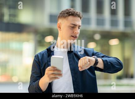 Busy young man with curly hair checking time on his smartwatch while holding a smartphone Stock Photo