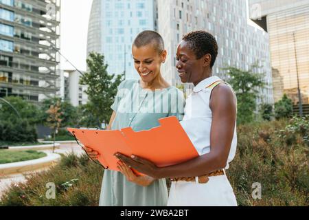Two Professional Women Examining Documents Together in an Urban Park During a Sunny Afternoon Stock Photo