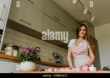 Portrait of friendly smiling female professional confectioner topping a cupcake with cream using a pastry bag. Looking at the camera. Indoors image. P Stock Photo