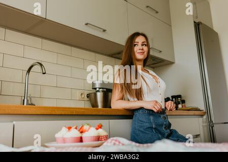 Portrait of friendly smiling female professional confectioner topping a cupcake with cream using a pastry bag. Looking at the camera. Indoors image. P Stock Photo