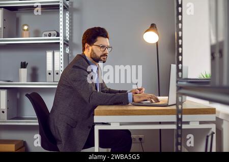 Side view of focused young, handsome man in glasses, dressed in office style, sitting at desk in front of laptop in the office and typing text and Stock Photo