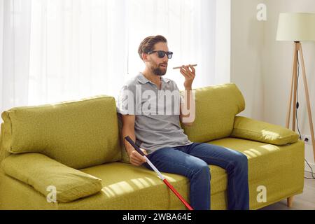 Blind man holding cane and using digital assistant on mobile phone sitting on sofa at home. Stock Photo